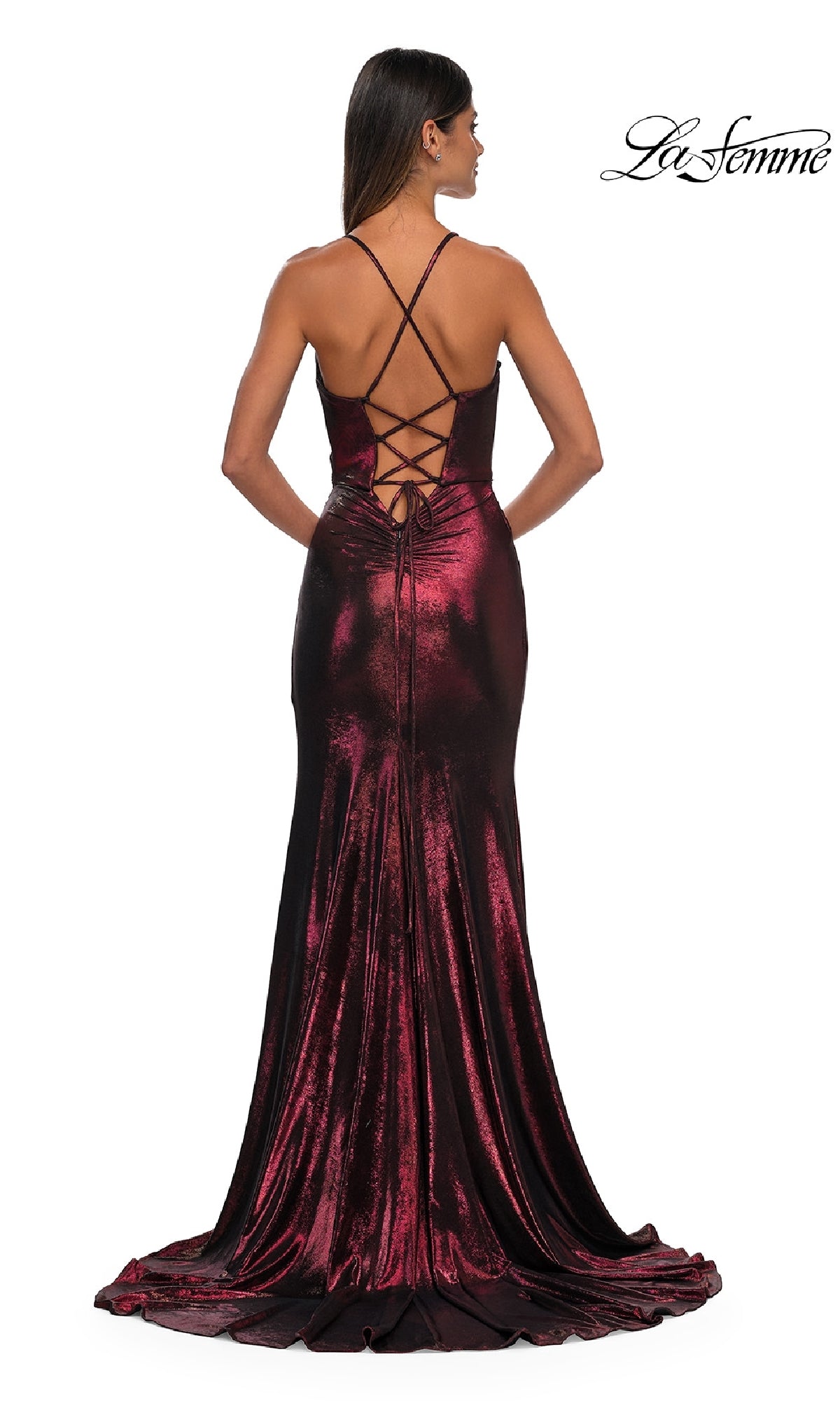 A woman in a deep red La Femme 32884 metallic prom dress with a lace-up open back and crisscross straps stands with her back to the camera, showcasing the gown’s long, flowing train for a dramatic effect.